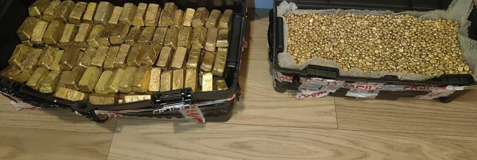 Deux mallettes ouvertes posées au sol : l'une remplie de lingots d'or brut coulés, l'autre de pépites et de petits lingots.